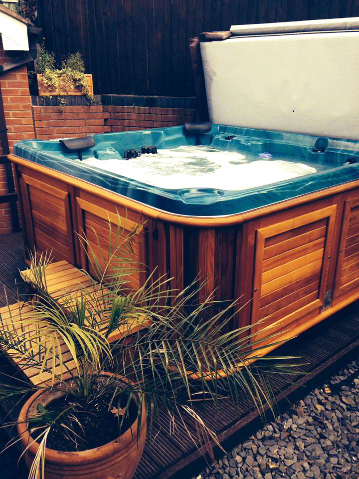 Wood‑frame hot tub on a deck with brick and fence backdrop, showcasing cozy hot tubs in Topeka.